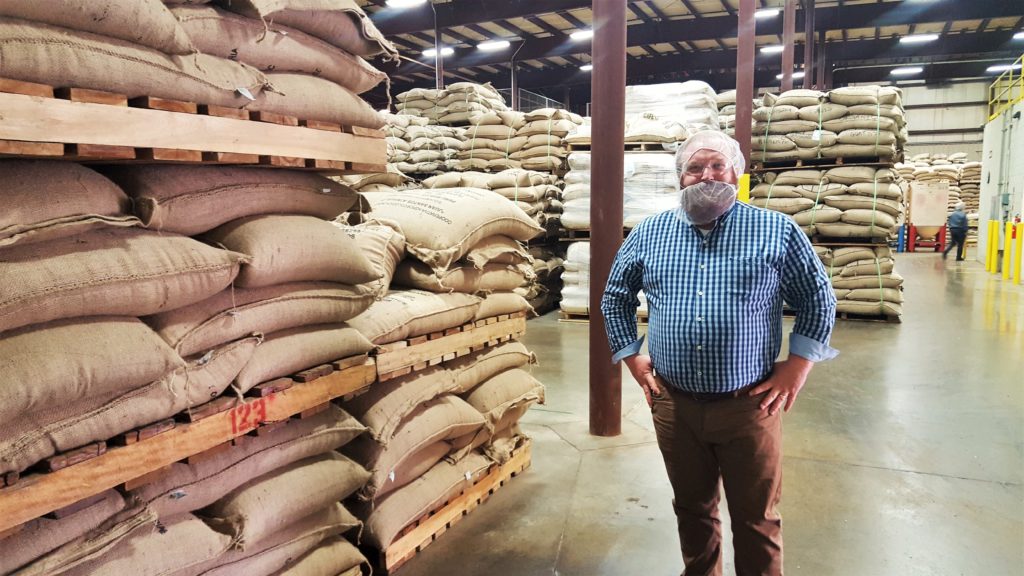 Mike Drescher at the entrance to the Paul deLima roasting facility. The stacks of burlap sacks are green coffee beans from various coffee-producing countries awaiting processing.